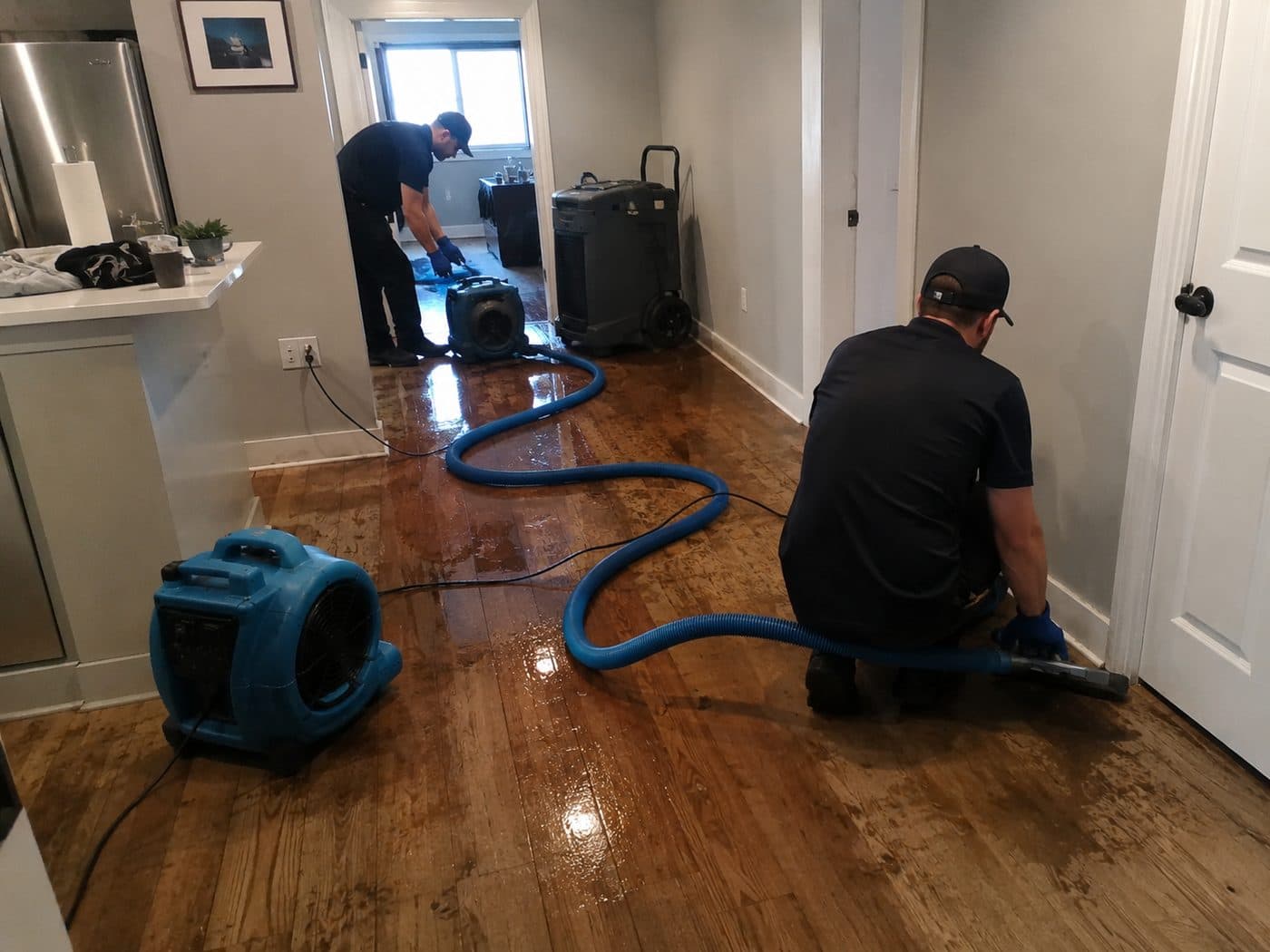 Two technicians extracting standing water from a hardwood floor with air movers running.