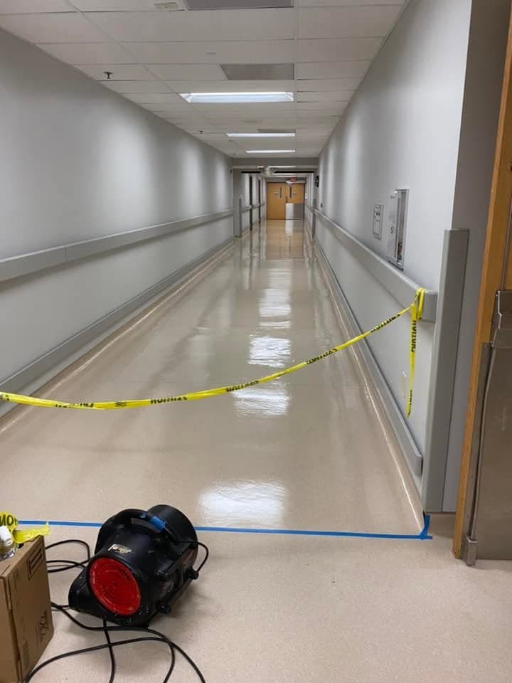 A freshly refinished hospital corridor floor drying behind caution tape, with an air mover running.