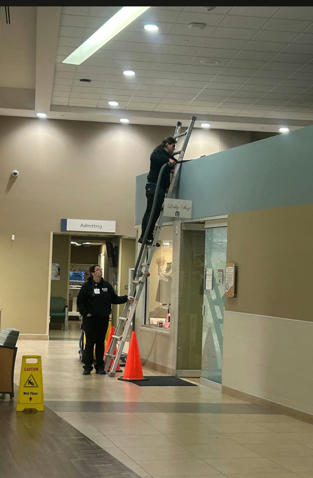 A G & J's technician on a ladder doing high level detail work in a hospital lobby while a floor machine runs below.