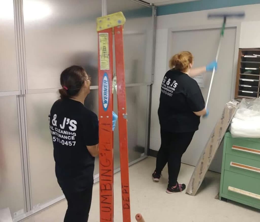 Two G & J's crew members cleaning an office interior, one working a door and partition, one steadying a ladder.