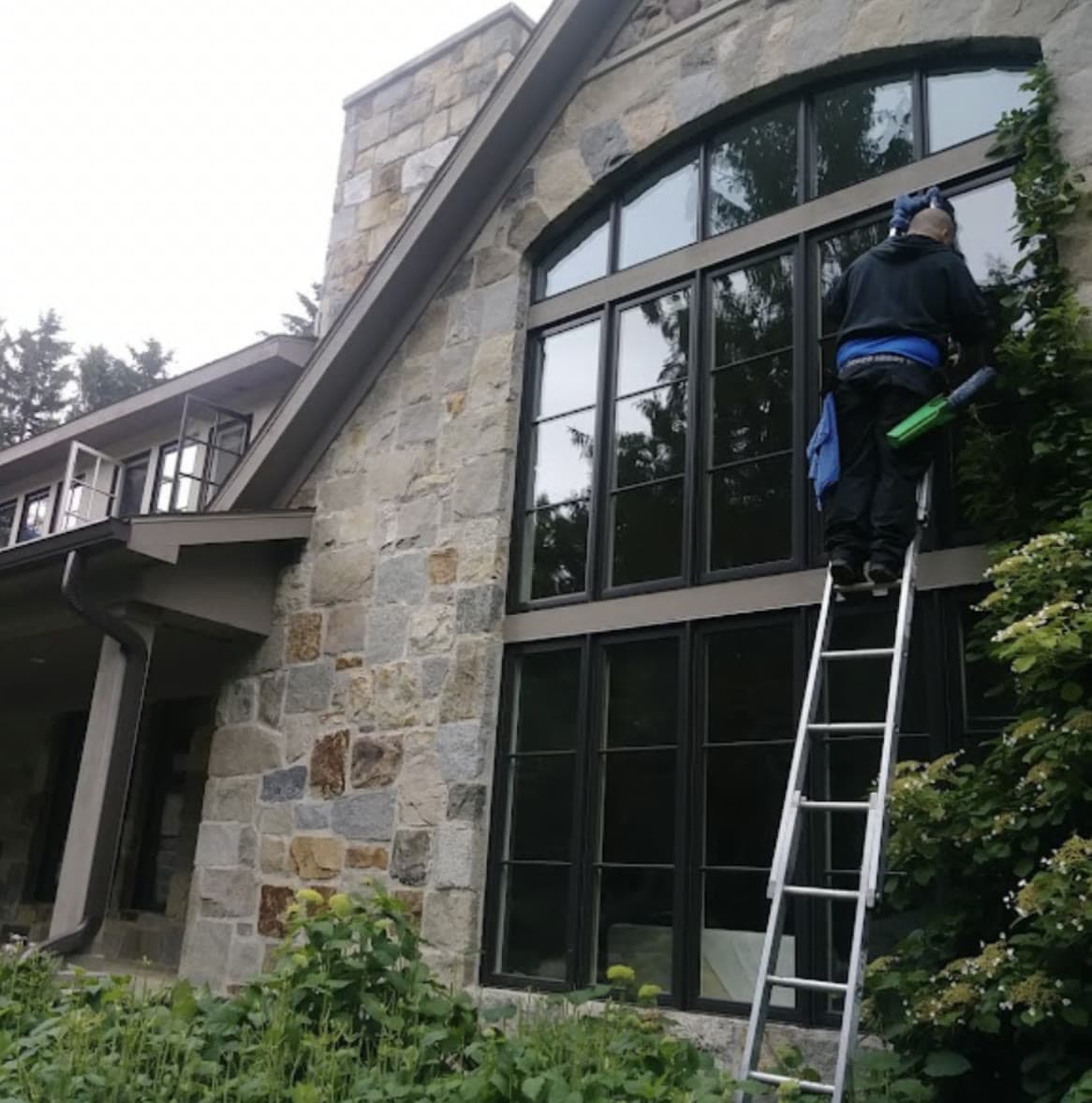 A G & J's technician on a ladder washing the tall windows of a stone house.